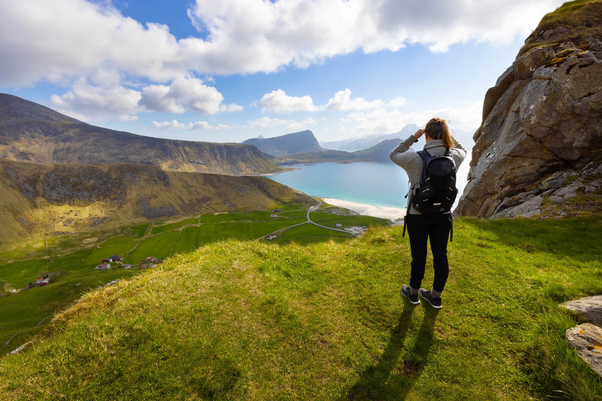 Person standing on a grassy cliff overlooking a scenic landscape with a lake and mountains.