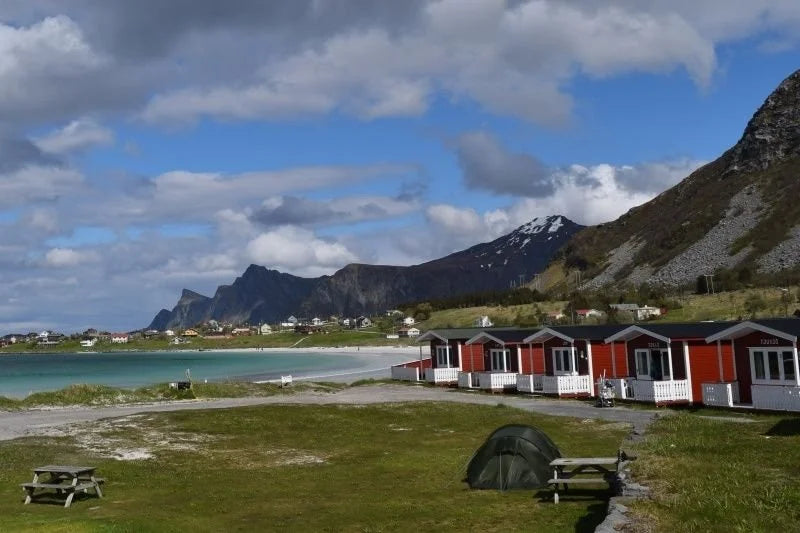 Scenic view of a campsite with cabins and a tent near a body of water with mountains in the background.