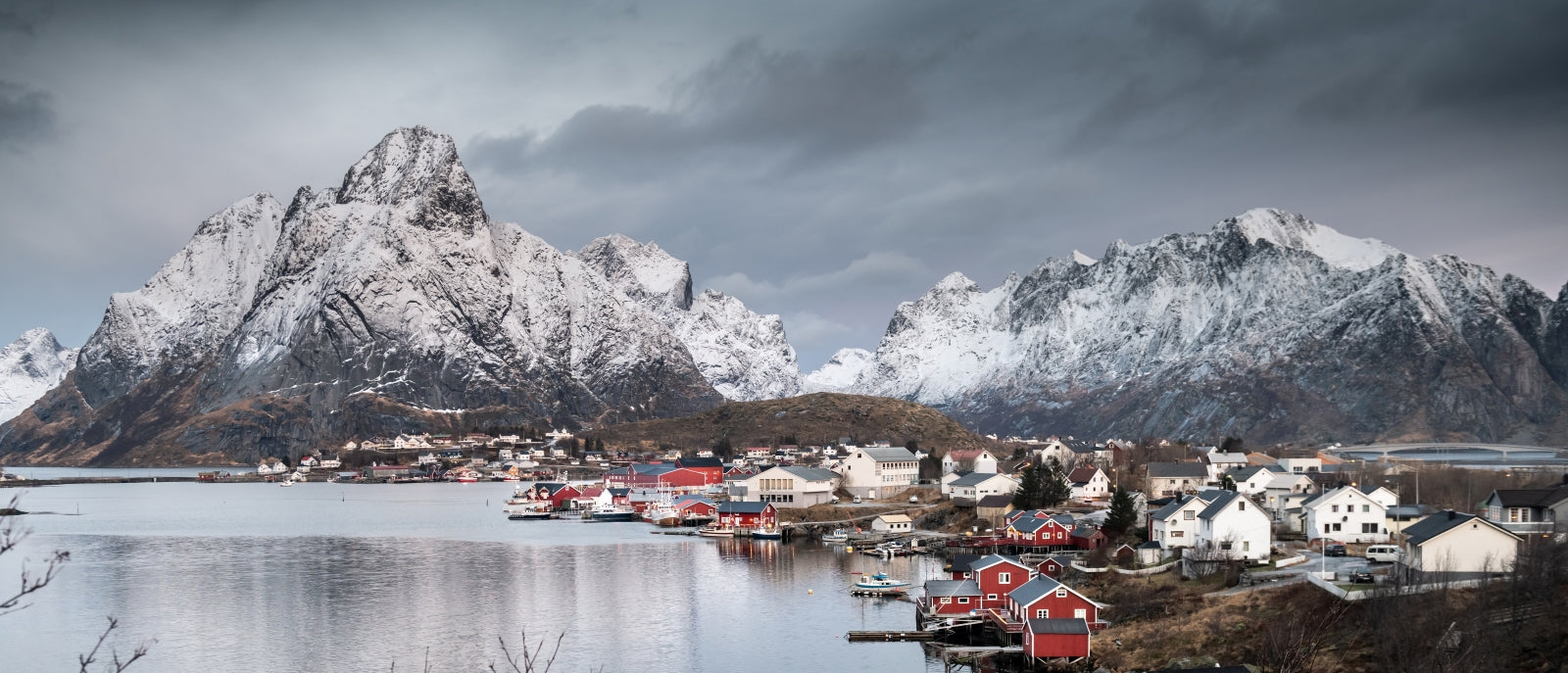 Scenic view of a coastal town with red buildings and snow-capped mountains under a cloudy sky.