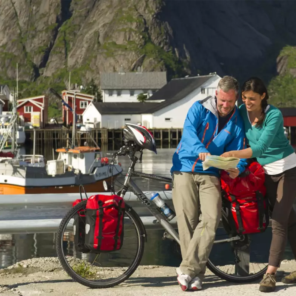Two people standing next to a bicycle with red bags, looking at a map by a waterfront with houses and mountains in the background.