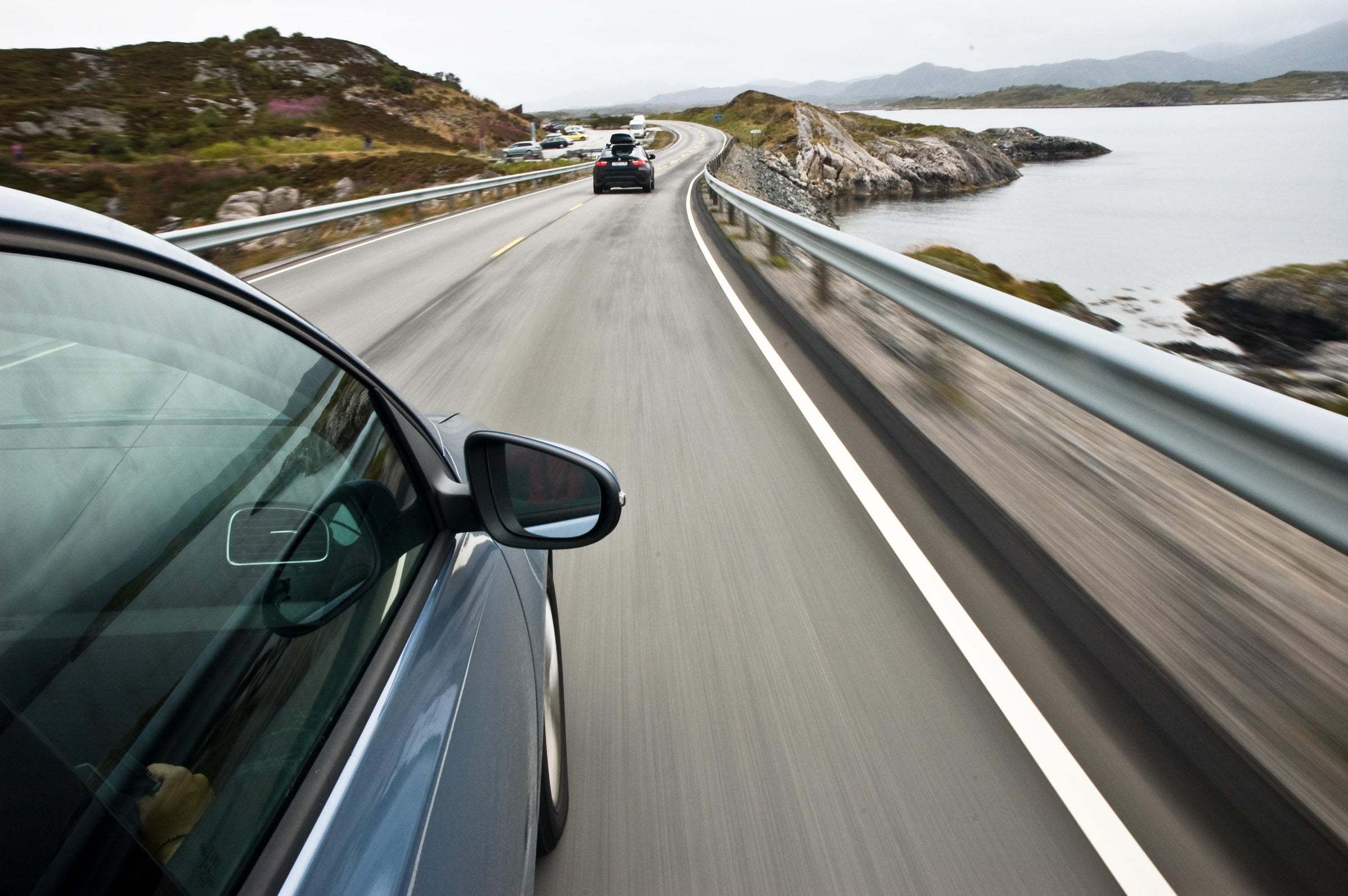 Car driving on a scenic coastal road with mountains and water in the background.