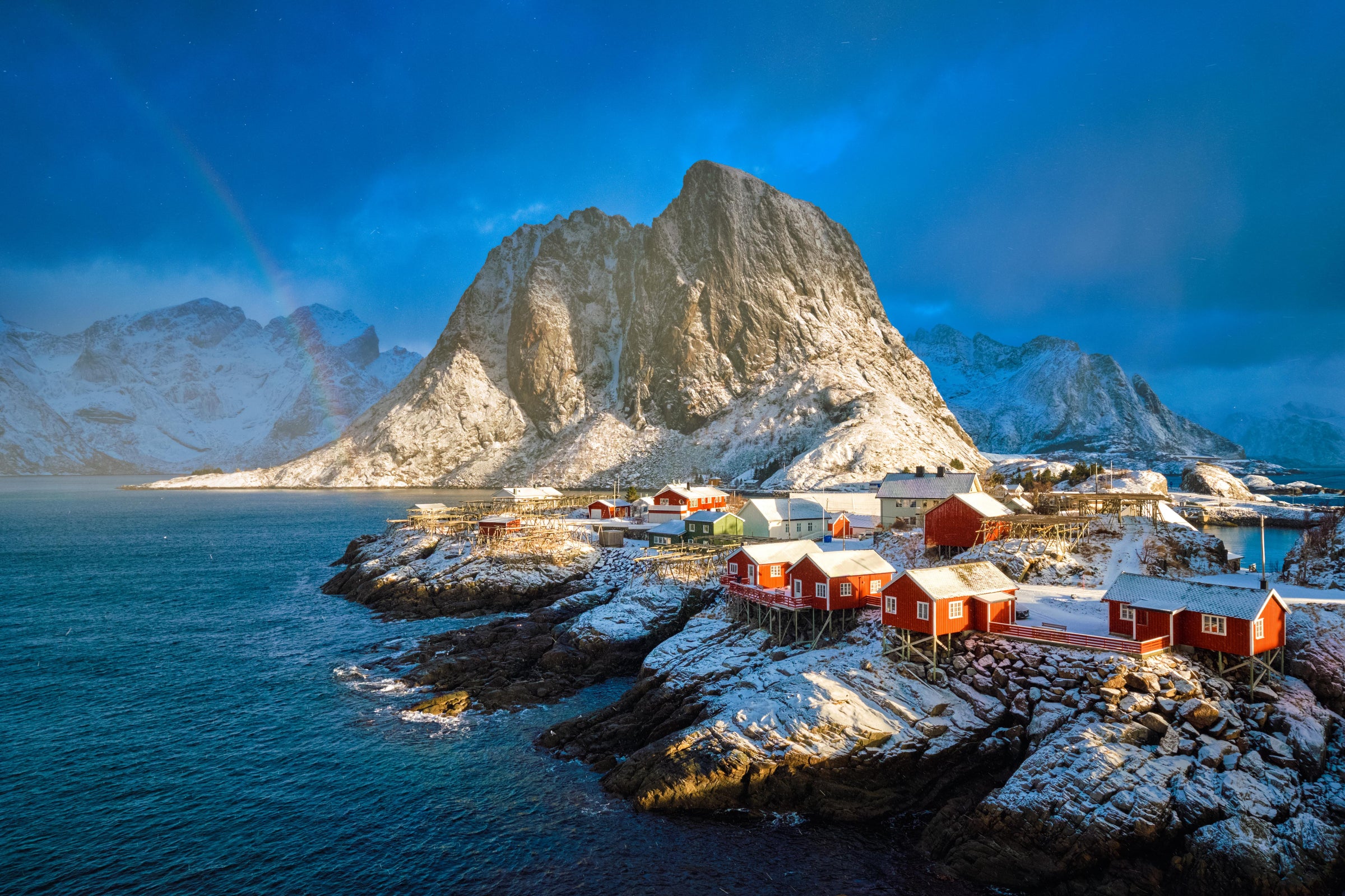 Red cabins on a rocky island with snow-capped mountains and blue sky.